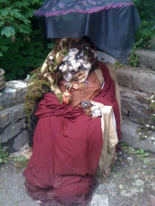 A life-size figure of a troll-like old woman, made out of felt, sitting in a chair.