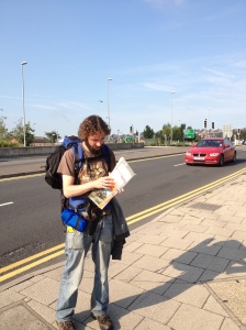Ross trying to look at an OS map, by a busy road, and looking confused. Ross has a beard and dark curly hair and is wearing a rucksack.